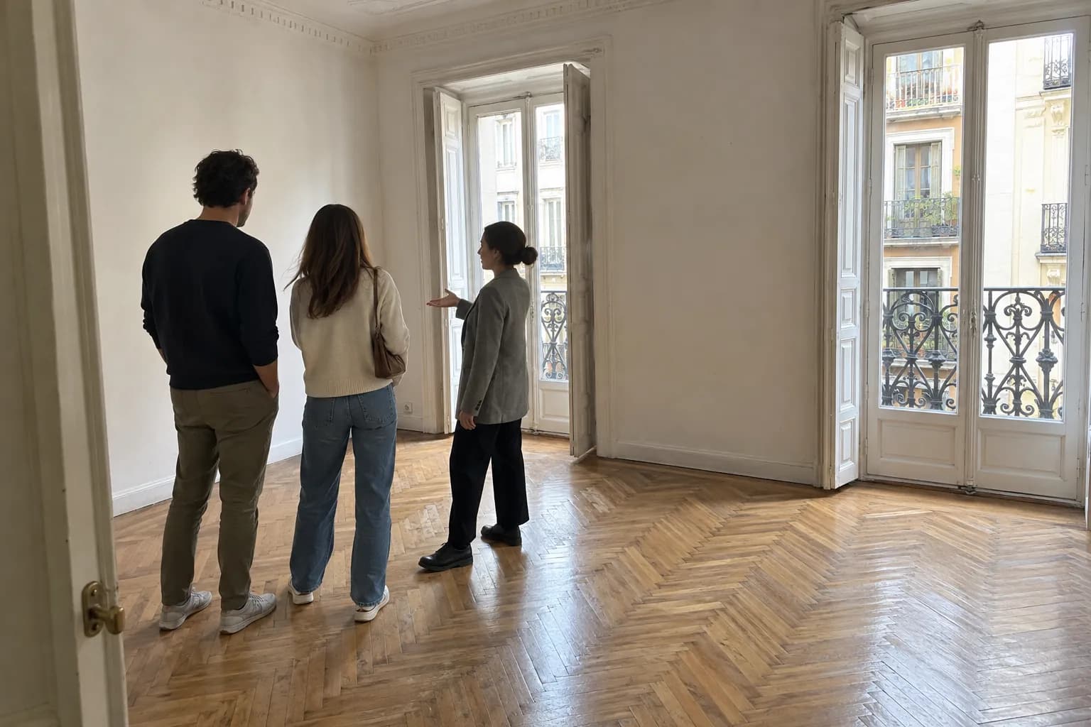 Una pareja visita un piso luminoso con balcones de forja acompañada por una asesora
